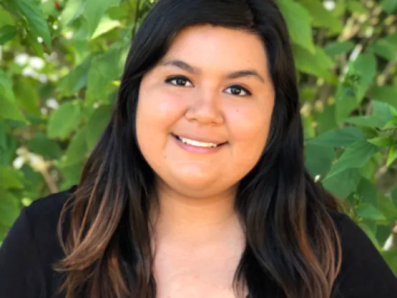 Bianca Aguilar, smiling Latina woman with long dark hair, with green foliage in background