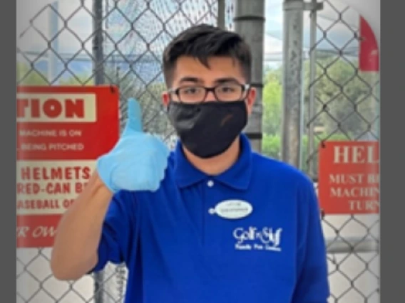 Young adult with short dark hair and glasses, wearing a blue shirt and a black medical mask, giving a thumbs-up to the camera.