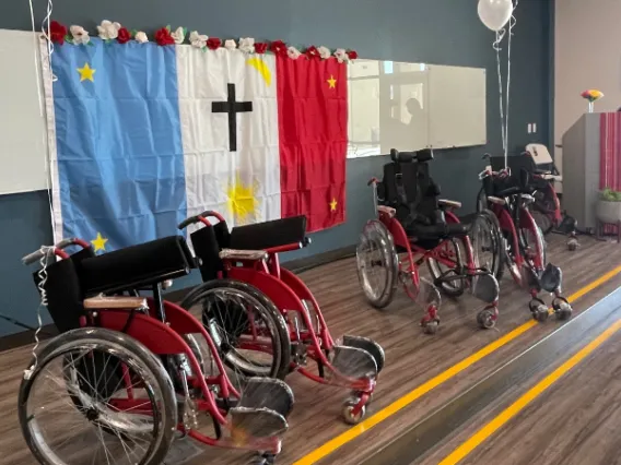 Several new red and black wheelchairs, still wrapped in cellophane, positioned side-by-side on a stage with a Tribal flag hung in the background.