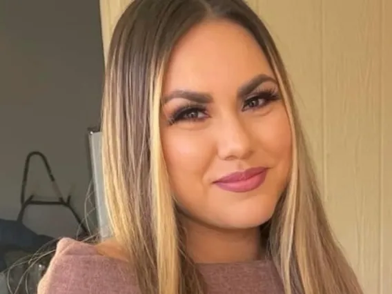 Alexis, a woman of color with long brunette hair with blonde highlights, is standing in front of a white background. She wears a brown sweater and smiles at the camera. 