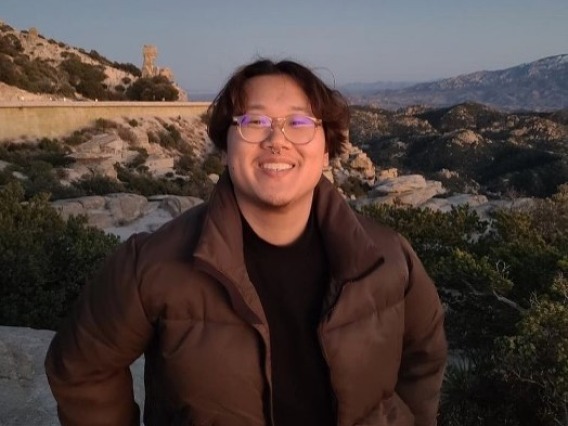 Andrew, a Chinese American man with dark brown hair, is outside in front of a mountain range. He wears clear-rimmed glasses and smiles at the camera. He also wears a black mock-neck and a brown puffer jacket. 