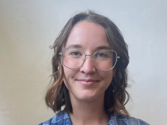 Audrey, a white woman with shoulder length brunette hair, standing in front of a white background. She wears silver rimmed glasses and smiles at the camera. She is also wearing a  blue checkered collared shirt. 