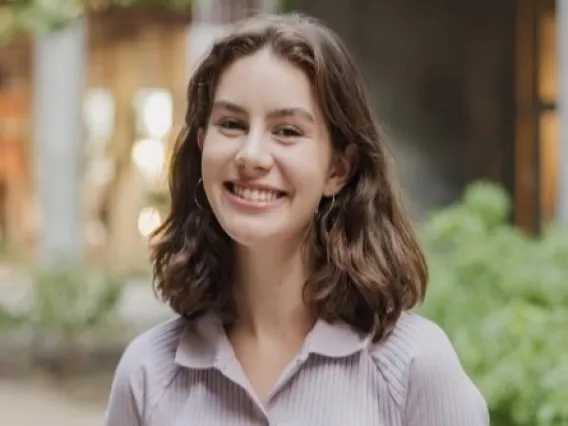 Cate has shouldner length burnete hair and is smiling at the camera, wearing a tan collared button up shirt and blue jeans.