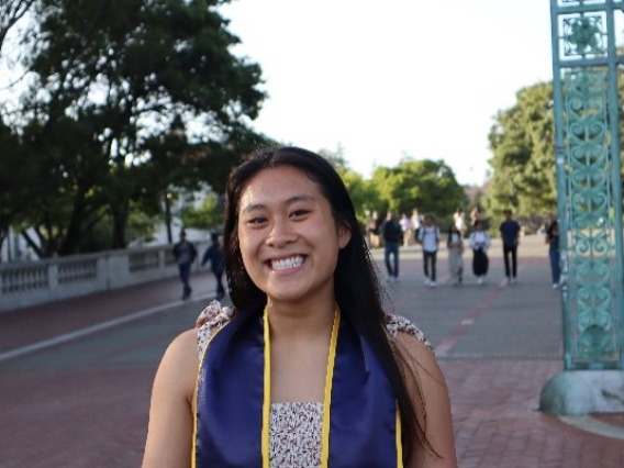 Cosette, an Asian woman with long black hair, smiling at the camera and standing outside in front of a big teal arch gate.  She is wearing a white dress with floral designs with a navy blue graduation stole with gold outlines. 