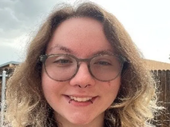 Elexander, a white man with shoulder-length brown hair with the ends bleached, is smiling at the camera. He has round green glasses, a black septum ring and two black lip rings. He is also wearing a black button-up shirt with brown and white leaves on it