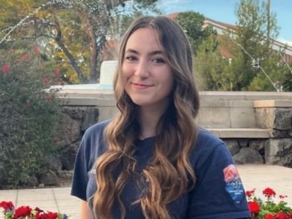 Ella, a white woman with curled, long brown hair, is outside on a bright day. She is sitting in front of a fountain and flowers, with trees in the background. She smiles at the camera and wears a navy blue short-sleeved T-shirt.