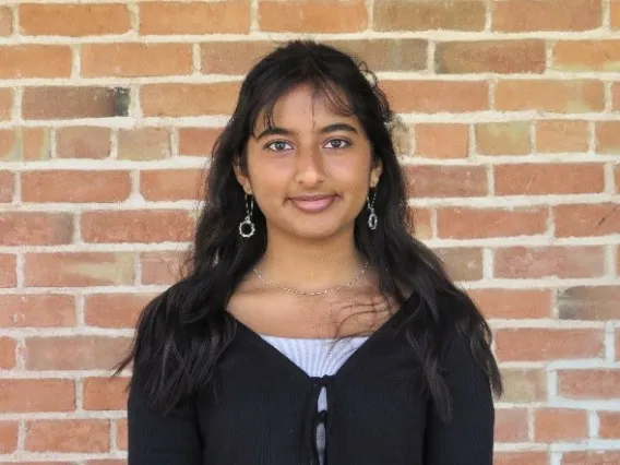 Esha has long black hair with bangs, is wearing a black long sleeve with a white under shirt is smiling at the camera in front of a brick background. 