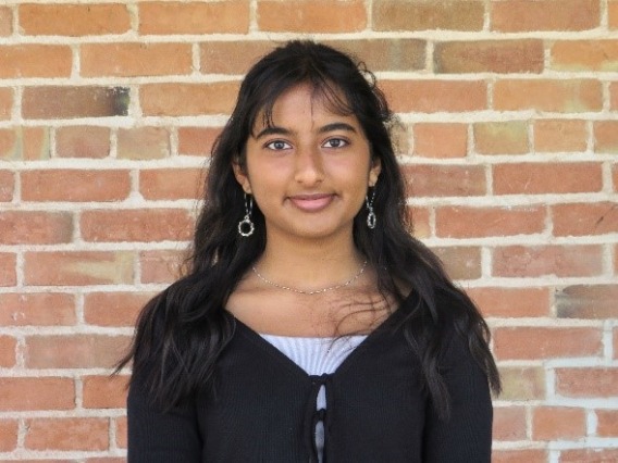 Esha has long black hair with bangs, is wearing a black long sleeve with a white under shirt is smiling at the camera in front of a brick background. 