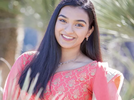 Freya, a woman of color with long black hair, is outside in front of a palm tree. She smiles at the camera and wears a traditional Indian dress in pink with gold detailing. 