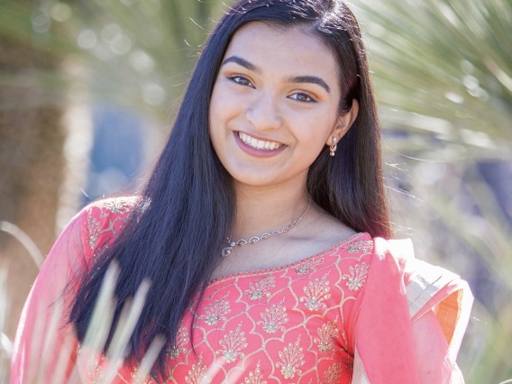 Freya, a woman of color with long black hair, is outside in front of a palm tree. She smiles at the camera and wears a traditional Indian dress in pink with gold detailing. 