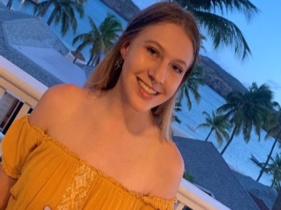 Gabby, a white woman with blonde hair and blue eyes is standing on a balcony in front of a blue backdrop of palm trees and the Caribbean ocean, wearing an off-shoulder yellow sundress.