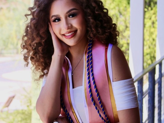 A young multi-ethnic woman with curly brown hair wearing a white dress and graduation stoles with desert landscape in the background.