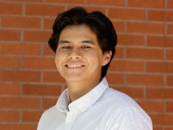 Joseph, a dark-skinned person with long brown hair, is outside on a bright day in front of a red brick wall. He wears a striped white shirt with black pants and a brown belt, and smiles at the camera.