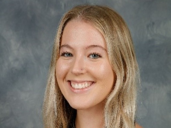 Kiley, a white woman with long blonde hair is standing in front of a grey background. She wears a black halter top and is smiling at the camera.