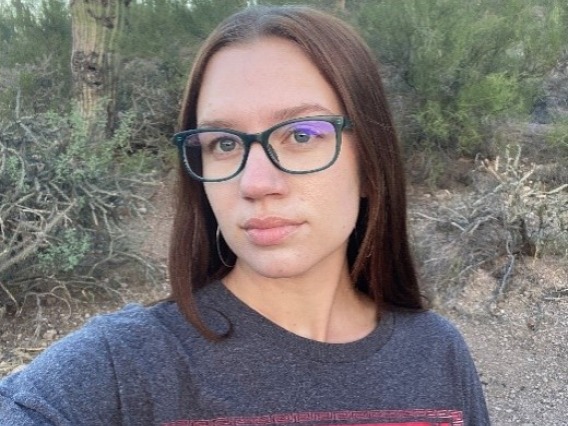 Kirsten, a white woman with red hair is outside. She is wearing green glass and a red and gray t-shit, standing in front of a desert landscape. 