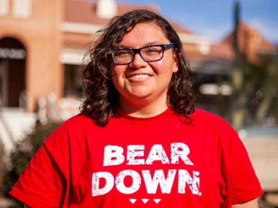 Lexicon, is facing the camera and smiling. His hands are behind his back and he stands in front of Old Main, which is blurred in the background. He is wearing a red shirt that says "BEAR DOWN" in all caps, and his hair is shoulder-length and curly. 