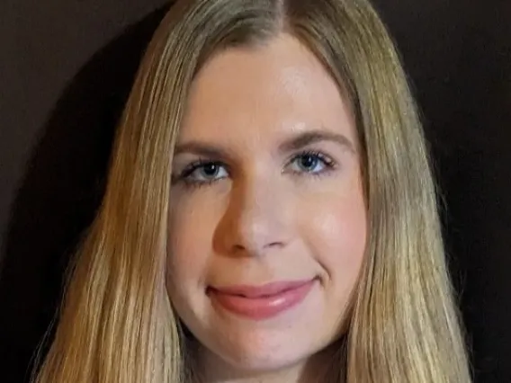 A white woman with blonde hair is smiling in front of a brown background. She is wearing a navy blouse that has white and copper geometric patterns. 