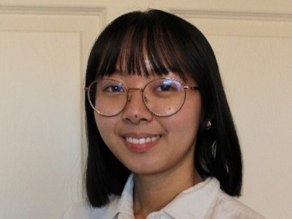 Nadine, an Asian person with shoulder-length black hair and bangs, is smiling in front of a white background. They are wearing gold-rimmed glasses and a white button-down shirt.