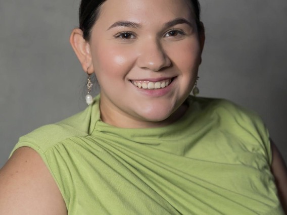 Paulette is sitting in front of a gray backdrop. She has dark hair and is wearing long pearl earrings with a lime green top. Paulette is smiling and looking at the camera.
