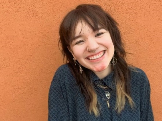 Ree, a Chinese American individual with blue and black checkered button up shirt. They are smiling in front of an orange wall outside on a bright day, wearing long alternative earrings, 3 necklaces, and their hands in their pockets.