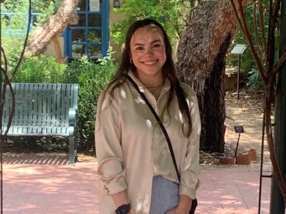 In this image, I am wearing a gold satin blouse with a denim skirt. My hair is down and I am also wearing a black small purse. The background of the image shows trees and plants from the Tucson Botanical Garden.