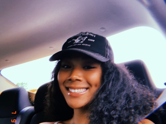 Brown skinned female with long curly hair selfie taken in the back of a car, wearing a black/white hat, white tank top, with a silver/gold necklace, smiling at the camera