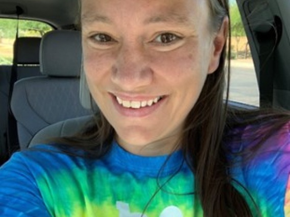 Katie Kwiatkowski, a woman with long brown hair, wearing a tie-dye shirt, smiling and sitting in a car