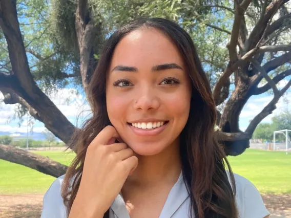 Aaliyah Washington, a young woman with long brown hair, wearing a light blue top, standing outside, in front of a tree, smiling