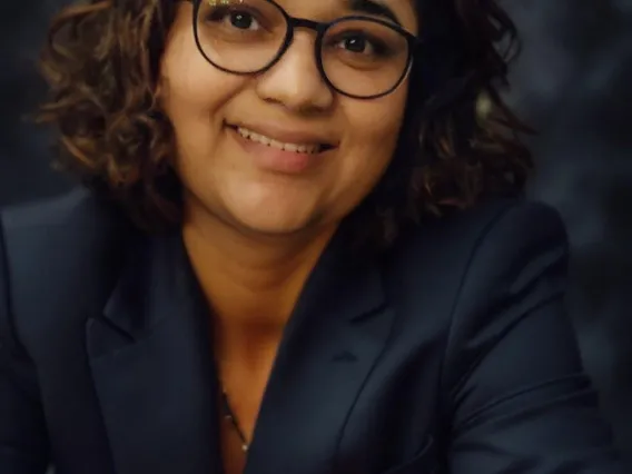 Woman with short, curly dark hair wearing glasses and a dark grey blazer.