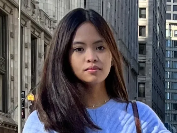 Dana has shoulder length brunette hair, standing in front of buildings wearing blue sweater look at the camera. 