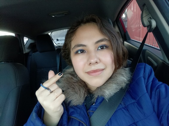 Leslie, a light-skinned woman, with short brown hair, and brown eyes, is sitting inside a car. She is wearing a blue puffer jacket, a blue ring on her right index finger, and a black ring on her middle finger, her nails have black nail polish, and she is wearing blue eyeshadow. She is smiling lightly. 