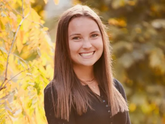 Samantha Zwillinger, a young woman with long brown hair, wearing a brown long sleeve top, standing outside among trees with yellow leaves, smiling