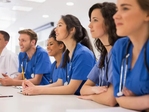 two young men and four young women sit at a table and look ahead. They are wearing blue scrubs with stethoscopes around their necks. 