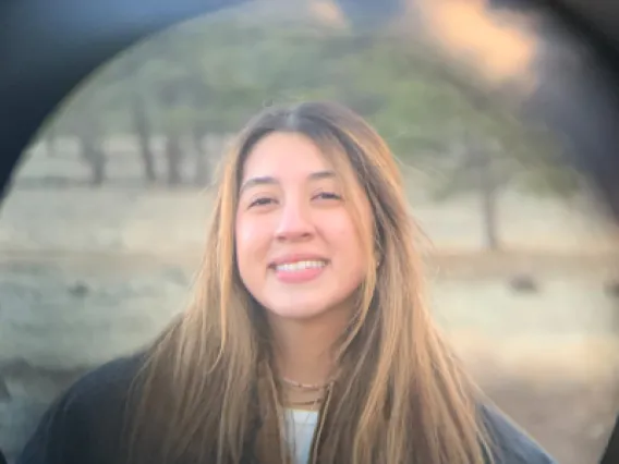 Anahy, a hispanic woman with long brown hair, is outside. She wears a black jacket and smiles at the camera.