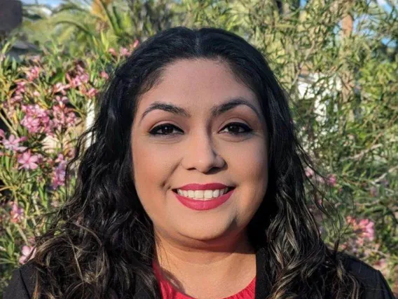 young woman smiling at camera, there are cacti and flowers in the distance. She is wearing a red shirt with a black blazer and a University of Arizona logo pinned on top. 