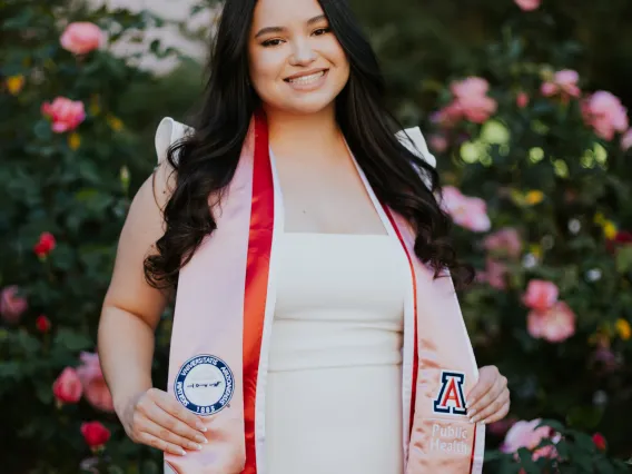 Photo of young woman in her University of Arizona cap and gown, long black hair smiling at camera. Standing in front of rose bushes