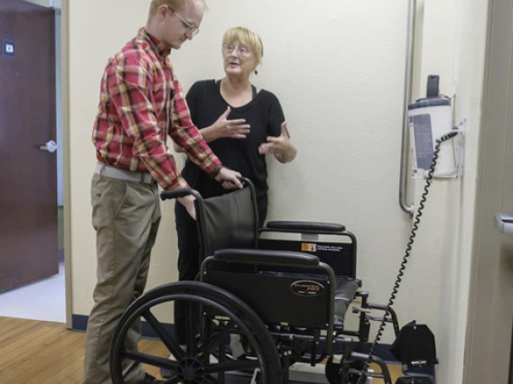 a gentleman is standing behind a wheelchair and holding onto its handles while a woman gestures with her hands beside him. The wheelchair is on a scale.