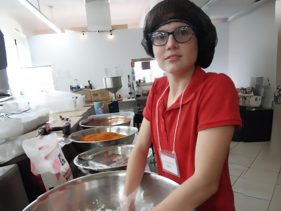Student wearing a hairnet and gloves hand mixing food in a large commercial mixing bowl.