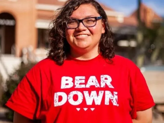 Lexicon Espinoza, a UA student with shoulder-length brown hair and glasses, wearing a Bear Down shirt, standing outside on the UA campus