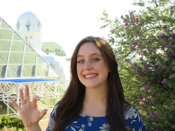 Nicole, a white woman with long brown hair, is outside on a bright day. The background includes a green bush with small purple flowers, and mountains in the background. She leans her side against a railing and smiles at the camera. She wears a royal blue blouse with white flowers, and black dress pants.