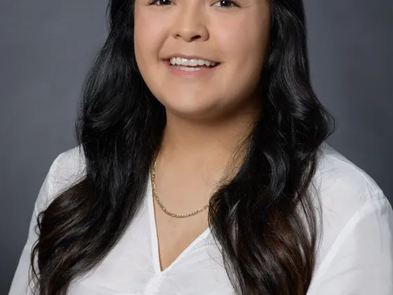 A young woman in front of a grey backdrop, with long brunette hair and wearing a white button down shirt smiling at the camera