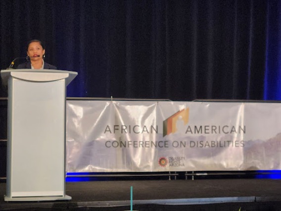 Jacy Farkas, speaking at a podium, with a banner in the background that reads "African American Conference on Disabilities - Disability Rights Arizona" with the AACD logo.