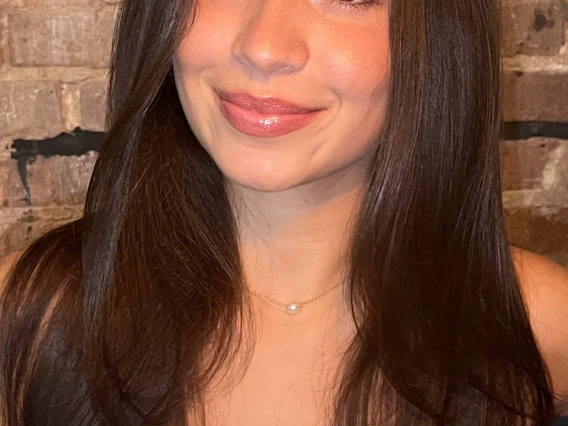 Woman with long brunette hair in front of brick background, smiling at the camera, wearing a white shirt