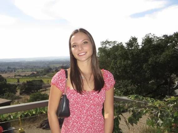 Phot of woman standing in front of green landscape, wearing a pink floral dress with brunette hair is smiling at the camera