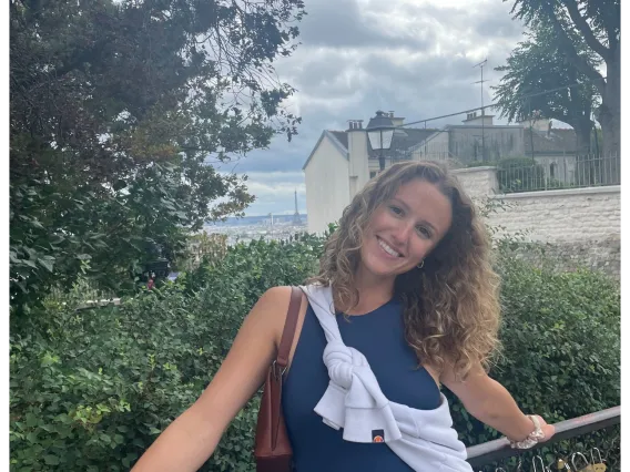 Photo of woman in front of a grassy landscape, wearing a navy blue top and white skirt, with blonde curly hair, smiling at the camera