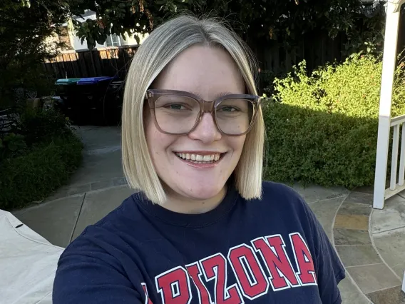 Photo of woman in front of greenery, wearing a University of Arizona shirt. She has a blonde bob and is wearing glasses