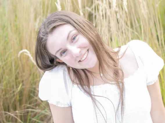 Photo of young woman in front of a field, wearing a white shirt with long brunette hair smiling at the camera