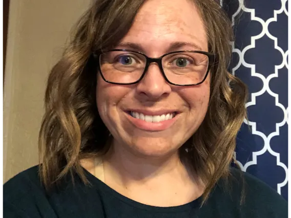 Photo of woman with brunette shoulder length hair wearing a dark blue shirt and black rimmed glasses, and she is smiling at the camera