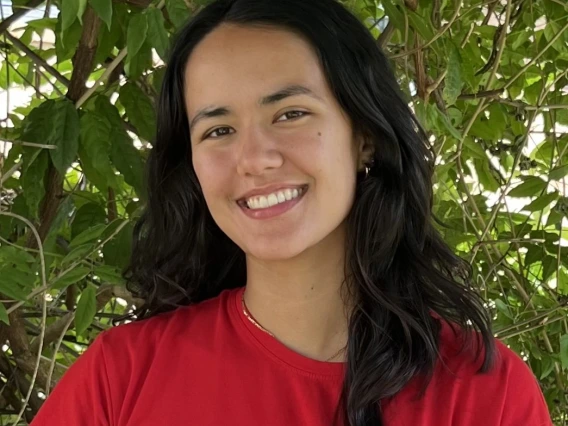 In the center of this image is me, Jeciana, an individual with light colored skin and dark black hair that falls just below my shoulder. In the picture, I am wearing a red shirt and am smiling in front of a large green plant.