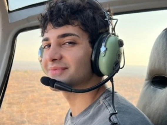 Photo of young man wearing a blue grey long sleeve shirt, has pilot headphone on and is in a helicopter. He has brown curly short hair and is smiling at the camera